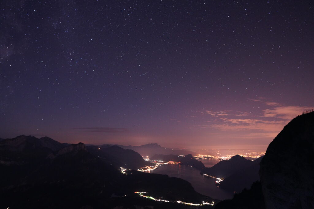 A colorful sunset view over Lake Lucerne from Fronalpstock, with glowing city lights illuminating the valley below.