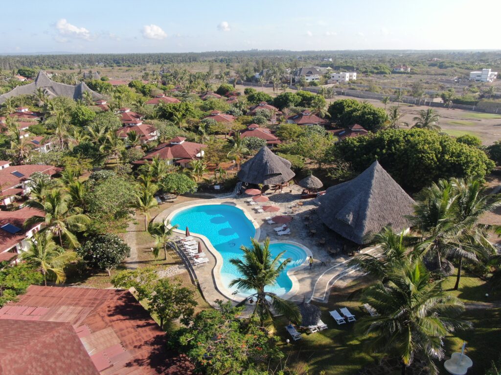 Aerial view of a beautiful beachfront resort in Watamu featuring traditional thatched roofs, lush palm trees, and a large swimming pool.